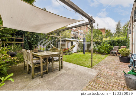 A serene garden retreat showcasing a dining table, hammock, and lounge chairs surrounded by lush greenery and a brick pathway. 123380059