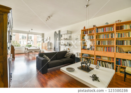 A stylish modern living room featuring a sleek black sofa, wooden shelves filled with books, and a bright ambiance created by natural light. 123380286