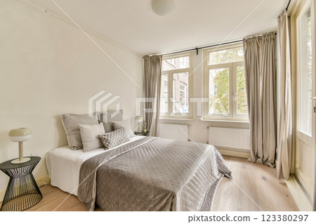 A stylish modern bedroom featuring a comfortable bed with light gray bedding, elegant curtains, and a chic decorative lamp. Bright natural light enhances the serene atmosphere. A stylish modern bedroom featuring a comfortable bed with light gray bedding, elegant curtains, and a chic decorative lamp. Bright natural light enhances the serene atmosphere. 123380297