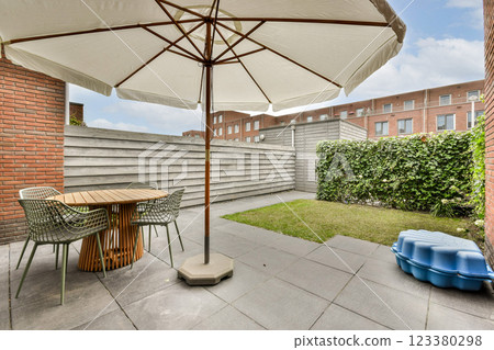An inviting outdoor patio showcases a round table with stylish chairs, a large umbrella, and a well-maintained green space, perfect for relaxation and gatherings. 123380298