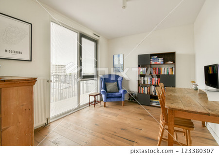 Bright and airy room featuring a blue chair, wooden flooring, and a bookshelf filled with books, creating a cozy reading nook. 123380307