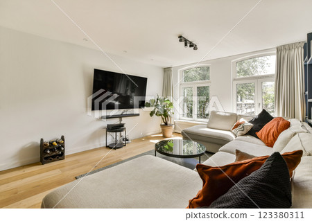 A stylish modern living room featuring a large TV, elegant seating, and ample natural light streaming through big windows, creating a cozy ambiance. 123380311