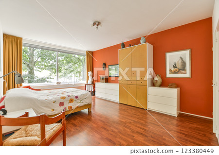 A stylish modern bedroom featuring wooden flooring, orange walls, large windows, and contemporary furniture arranged for comfort and aesthetics. 123380449