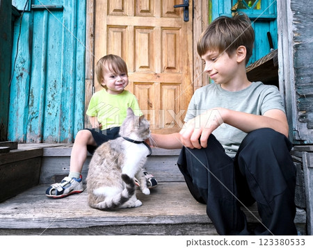 Two boys are sitting on the steps outside with a gray cat. A small child and his brother are cute petting a cat, children are playing with a cat outside. 123380533