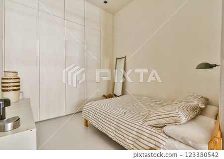 A serene and modern bedroom showcasing a simple design with a bed, striped bedding, a mirror, and clean lines, emphasizing tranquility and comfort. A serene and modern bedroom showcasing a simple design with a bed, striped bedding, a mirror, and clean lines, emphasizing tranquility and comfort. 123380542