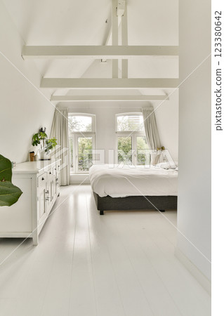 A spacious, light-filled bedroom with a minimalist design featuring white walls, large windows, and elegant furnishings that create a calm atmosphere. 123380642