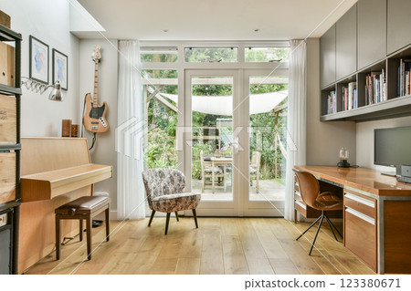 A modern home office interior featuring a wooden desk, chair, and musical instruments, complemented by natural light from a garden view. 123380671
