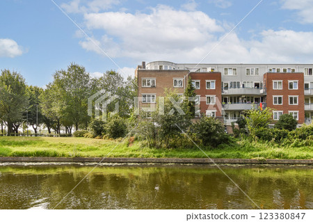 A picturesque view of a residential apartment building set against a backdrop of lush greenery and a serene water body, inviting tranquility and nature. 123380847