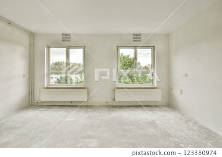 A spacious, unfurnished room featuring two large windows offering a view of greenery outside, enhancing the natural light in the space. 123380974