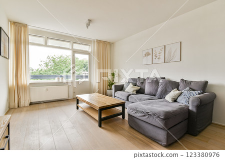 A stylish and inviting living room featuring a comfortable gray sofa, wooden coffee table, and large windows with natural light and greenery outside. 123380976