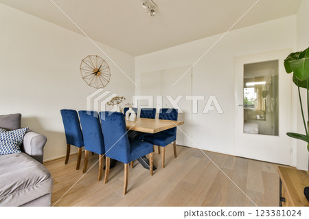 A contemporary living space featuring a dining table accompanied by stylish blue chairs, complemented by minimal decor and wooden flooring for a chic ambiance. 123381024