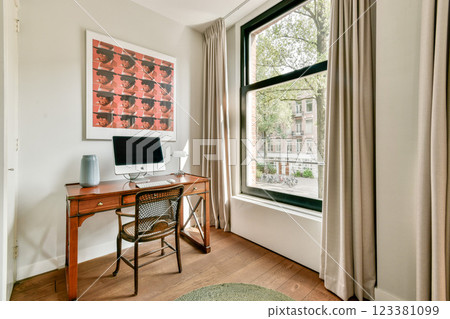 A stylish home office featuring a wooden desk, computer, and large window with curtains that lets in natural light, creating a serene workspace atmosphere. 123381099