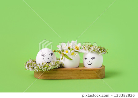 Three white eggs with a cheerful face and spring flowers of pears, strawberries and a white wreath of flowers on a green background. Happy Easter greeting card with a place for text. Three white eggs with a cheerful face and spring flowers of pears, strawberries and a white wreath of flowers on a green background. Happy Easter greeting card with a place for text. 123382926