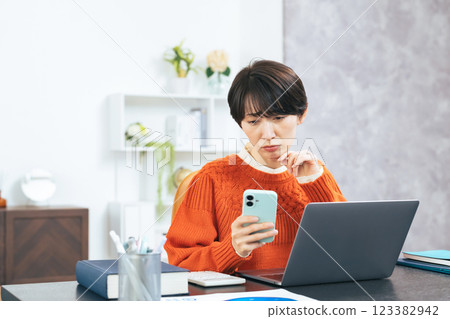 A middle-aged woman worried while looking at her smartphone and laptop A middle-aged woman worried while looking at her smartphone and laptop 123382942