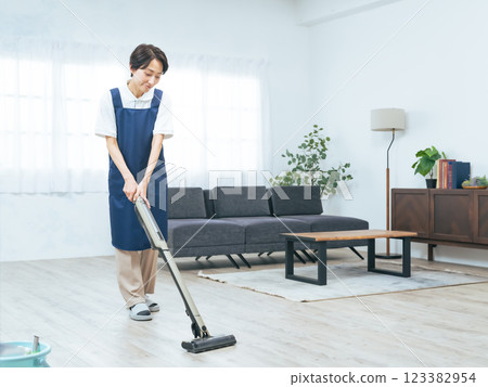 一名穿著圍裙的中年婦女正在用吸塵器清理房間的地板 一名穿著圍裙的中年婦女正在用吸塵器清理房間的地板 123382954