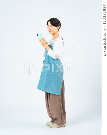 Full-body photo of a middle-aged woman wearing an apron using a smartphone Full-body photo of a middle-aged woman wearing an apron using a smartphone 123382987