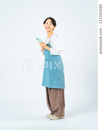 Full-body photo of a middle-aged woman wearing an apron using a smartphone 123382988