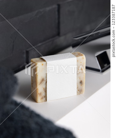 Beige soap with blank label on washbasin near faucet and dark grey bath towel close up, mockup 123387187