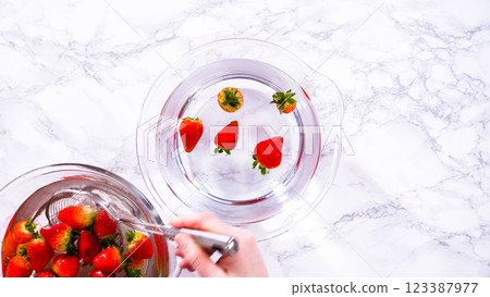 Flat lay. Ripe strawberries are submerged in water within a large glass mixing bowl, a step in washing the fruit to ensure cleanliness and longevity before storage or consumption. 123387977
