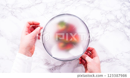 Flat lay. Strawberries, freshly washed and dried, are neatly stored in a glass bowl lined with a paper towel and securely covered with plastic wrap to maintain freshness. 123387986