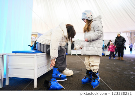 Mother helps daughter put on ice skates at winter skating rink in the afternoon Mother helps daughter put on ice skates at winter skating rink in the afternoon 123388281
