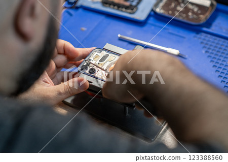 A skilled technician focuses on replacing a phone battery, surrounded by tools and equipment in a vibrant workshop setting. 123388560