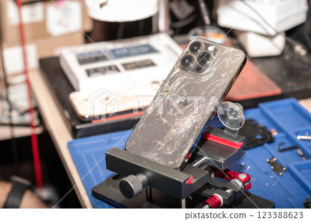 A damaged smartphone with broken glass is placed in a repair stand at a phone service shop, surrounded by tools and equipment. A damaged smartphone with broken glass is placed in a repair stand at a phone service shop, surrounded by tools and equipment. 123388623