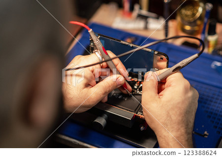 A technician is carefully repairing a smartphone battery with precision tools while working in a well-equipped workshop. A technician is carefully repairing a smartphone battery with precision tools while working in a well-equipped workshop. 123388624