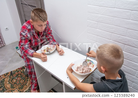Two young children are seated at a table, enjoying bowls of cereal. One child is wearing pajamas, and the other is in a dark shirt. Two young children are seated at a table, enjoying bowls of cereal. One child is wearing pajamas, and the other is in a dark shirt. 123389240