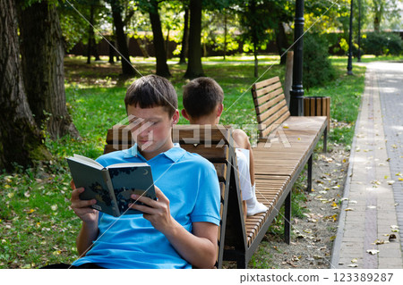 Two boys enjoying a sunny day. One engrossed in a book while the other is seated facing away enjoying the park scenery. Two boys enjoying a sunny day. One engrossed in a book while the other is seated facing away enjoying the park scenery. 123389287
