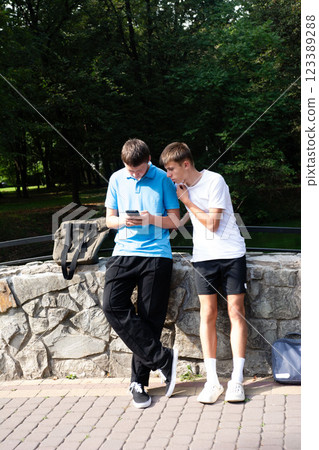 Two young people are looking at a cellphone together while standing by a rock wall with lush green trees in the background. Two young people are looking at a cellphone together while standing by a rock wall with lush green trees in the background. 123389288