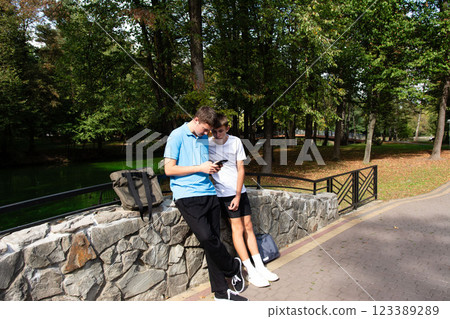 Two friends looking at a phone together while leaning against a stone wall in a park with a grassy area and trees in the background. Two friends looking at a phone together while leaning against a stone wall in a park with a grassy area and trees in the background. 123389289