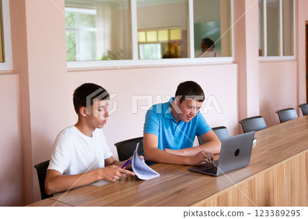 Two young men collaborate at a long table, one with a laptop and the other with papers, in a brightly lit room. Focus on teamwork. 123389295