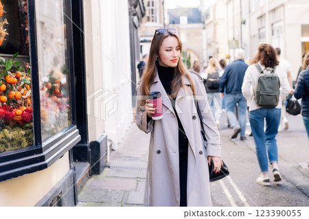 Young smiling woman in trench coat enjoying hot coffee drink in reusable cup while walking on the city streets decorated for seasonal holidays. Spring cozy mood, seasonal fashion style trend. Young smiling woman in trench coat enjoying hot coffee drink in reusable cup while walking on the city streets decorated for seasonal holidays. Spring cozy mood, seasonal fashion style trend. 123390055