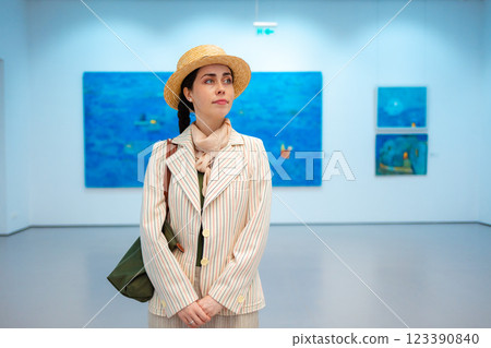 Mid shot of young Caucasian woman in elegant jacket, straw hat contemplating in art gallery at excursion. Big blue paintings in background. Concept of cultural education for students and Museum Day 123390840