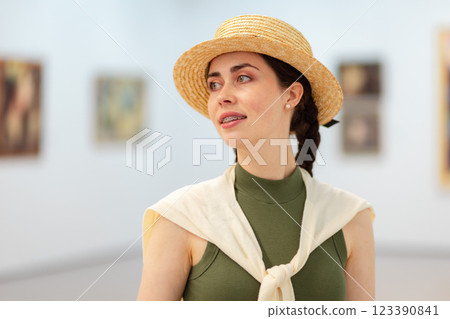 Portrait of a young Caucasian elegant smiling woman in straw hat posing in the art gallery. Paintings in background. Concept of cultural education and Museum Day 123390841