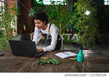 African woman florist gardener working in botanical store using laptop order plants communicating on Internet with customer. Female small business owner working at flower shop. Running of own business African woman florist gardener working in botanical store using laptop order plants communicating on Internet with customer. Female small business owner working at flower shop. Running of own business 123392526