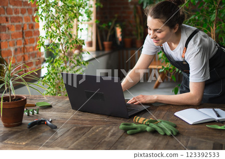 Female florist gardener working in botanical store greenhouse using laptop order plants communicating on Internet with customer. Small business owner working at flower shop. Running of own business Female florist gardener working in botanical store greenhouse using laptop order plants communicating on Internet with customer. Small business owner working at flower shop. Running of own business 123392533