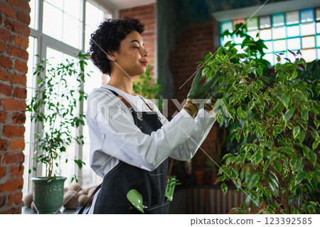 Running of own business. African woman florist wearing apron taking care of plants in botanical store. Happy small business owner working at flower shop smiling surrounded by plants Small business 123392585