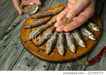 Hands expertly sprinkle spices or coriander over a line of fresh fish arranged on a wooden board. The rustic kitchen ambiance adds warmth, with vibrant colors from spices enhancing the scene 123393713