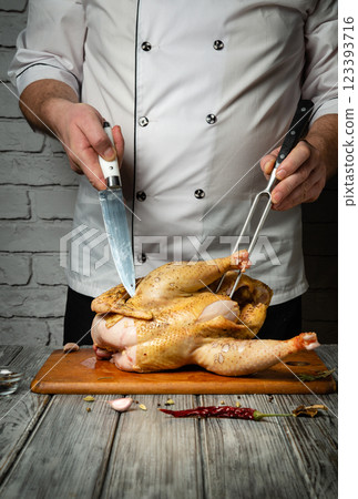 A skilled chef focuses intently while carving a raw chicken on a wooden board, surrounded by fresh herbs and spices. The warm glow of the kitchen enhances the culinary atmosphere 123393716