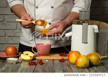 Making sweet apple juice by the hands of an experienced cook. Adding honey with a wooden spoon to a mug of fresh juice. Juice from apples in the kitchen using an electric juicer 123393717