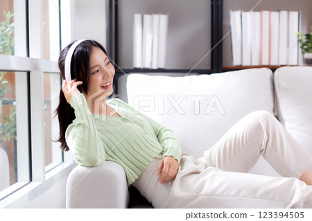 Happy young asian woman wearing headphones listening music while sitting on sofa with fun. 123394505