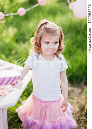 Happy birthday little girl making wish blowing candles on cake with pink decor in beautiful garden. 4 years birthday. child eat happy birthday pink cupcake. colorful pastel decoration outdoor 123398698