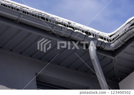 Modern metal gutter system with icicles and snow-covered eaves, highlighting winter conditions and roof drainage against a clear blue sky. 123400502