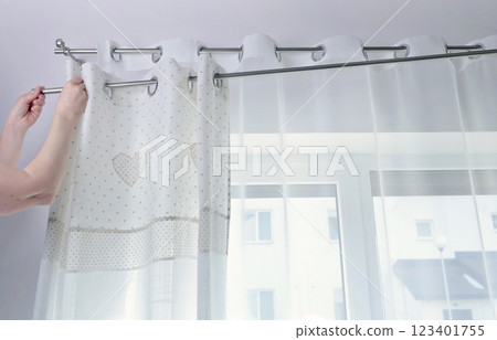 Woman hangs white curtains with metal rings made of natural material with embroidered hearts in kitchen of house. Cleaning and cleanliness. Interior and textiles. Woman hangs white curtains with metal rings made of natural material with embroidered hearts in kitchen of house. Cleaning and cleanliness. Interior and textiles. 123401755