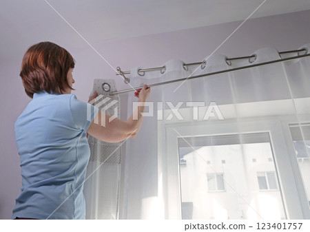 Woman hangs white curtains with metal rings made of natural material with embroidered hearts in kitchen of house. Cleaning and cleanliness. Interior and textiles.  123401757