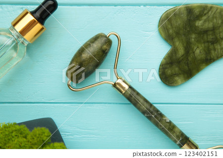 Green jade roller and gua sha stone for facial massage and on blue background. Home beauty and selfcare accessories. Face roller for anti age wrinkle treatment. Top view, flat lay. Green jade roller and gua sha stone for facial massage and on blue background. Home beauty and selfcare accessories. Face roller for anti age wrinkle treatment. Top view, flat lay. 123402151