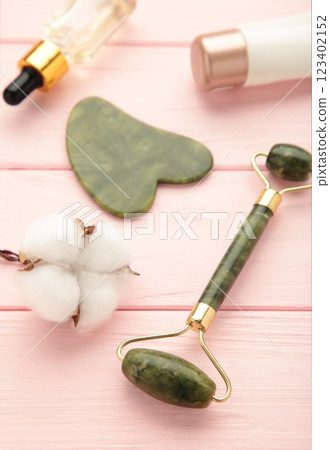Green jade roller and gua sha stone for facial massage and on pink background. Home beauty and selfcare accessories. Face roller for anti age wrinkle treatment. Top view, flat lay. 123402152