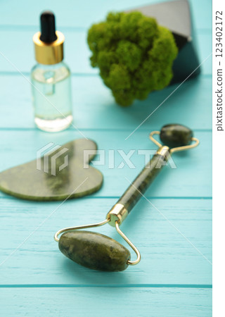 Green jade roller and gua sha stone for facial massage and on blue background. Home beauty and selfcare accessories. Face roller for anti age wrinkle treatment. Top view, flat lay. Green jade roller and gua sha stone for facial massage and on blue background. Home beauty and selfcare accessories. Face roller for anti age wrinkle treatment. Top view, flat lay. 123402172
